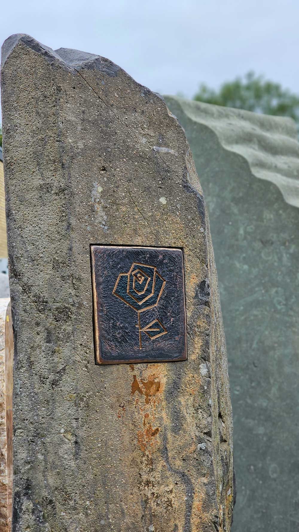 Nahaufnahme eines verwitterten Steins mit einer kleinen, quadratischen Plakette, die ein Rosenmotiv darstellt.