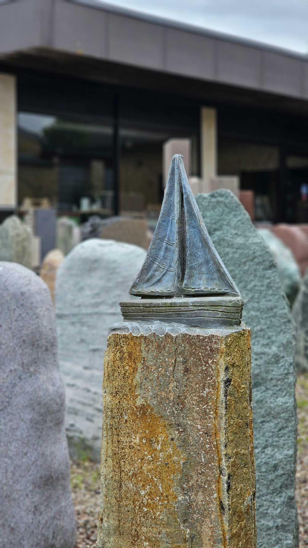 Bronzene Segelbootskulptur auf einer Steinsäule, umgeben von verschiedenen großen Steinen; im Hintergrund ein Gebäude.