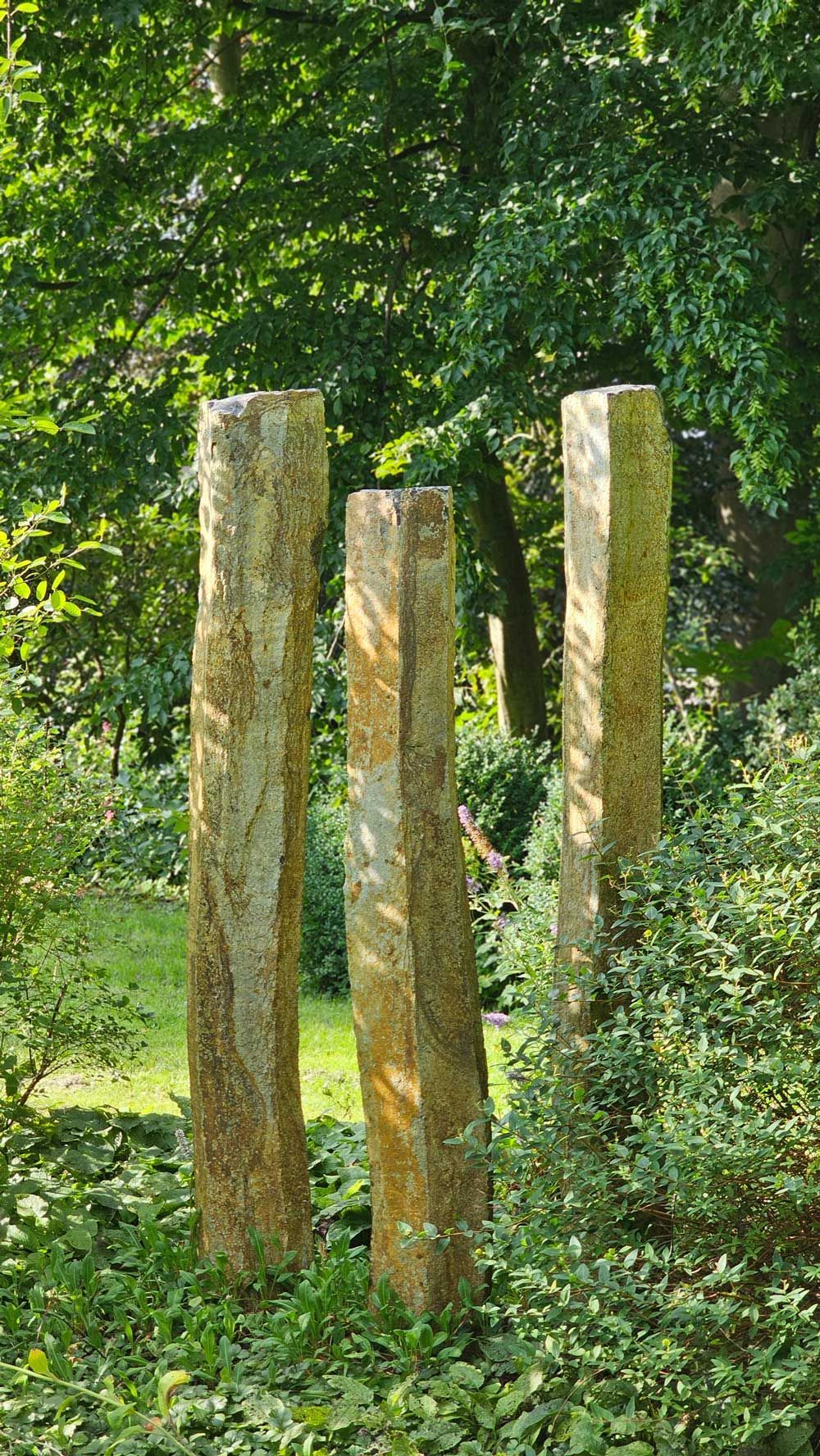 Drei hohe, verwitterte Steinsäulen in einer Gartenanlage, umgeben von üppigem Grün und Bäumen.