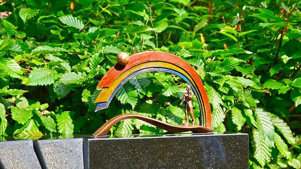Bronzeskulptur einer Person unter einem Regenbogenbogen vor grünem Laub.