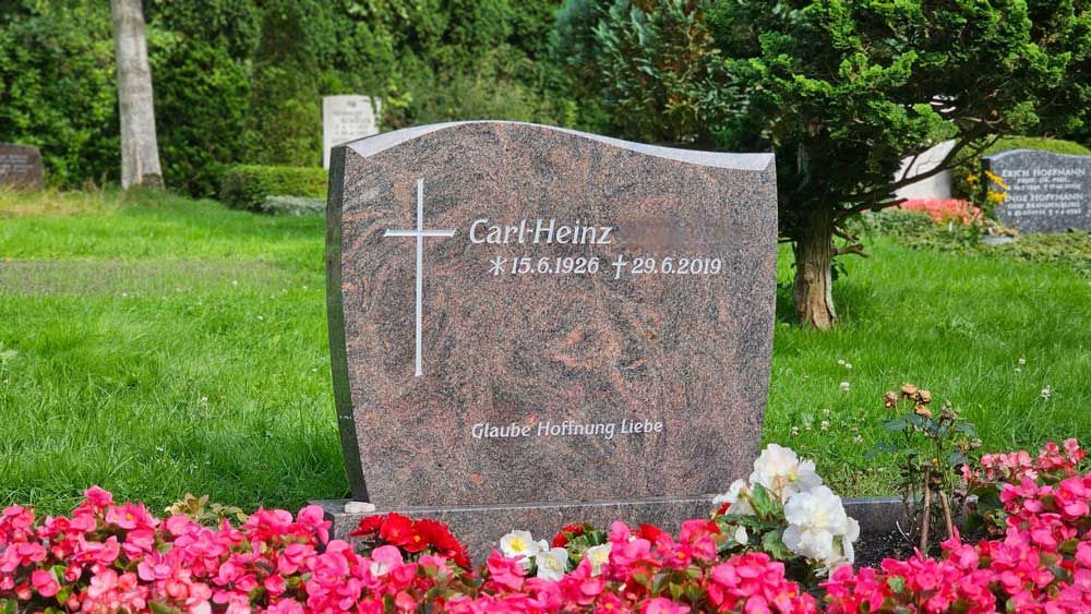 Grabstein mit Kreuz und Inschrift auf einem grünen Friedhof, umgeben von Blumen.