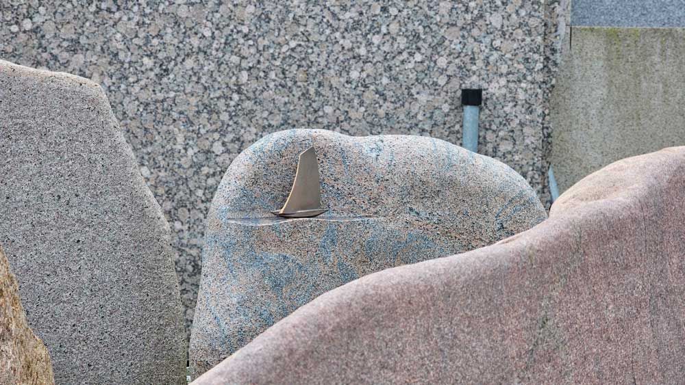 Große, glatte, mehrfarbige Steine, in einem davon befindet sich ein Metallgegenstand. Im Hintergrund eine graue Kiesmauer.