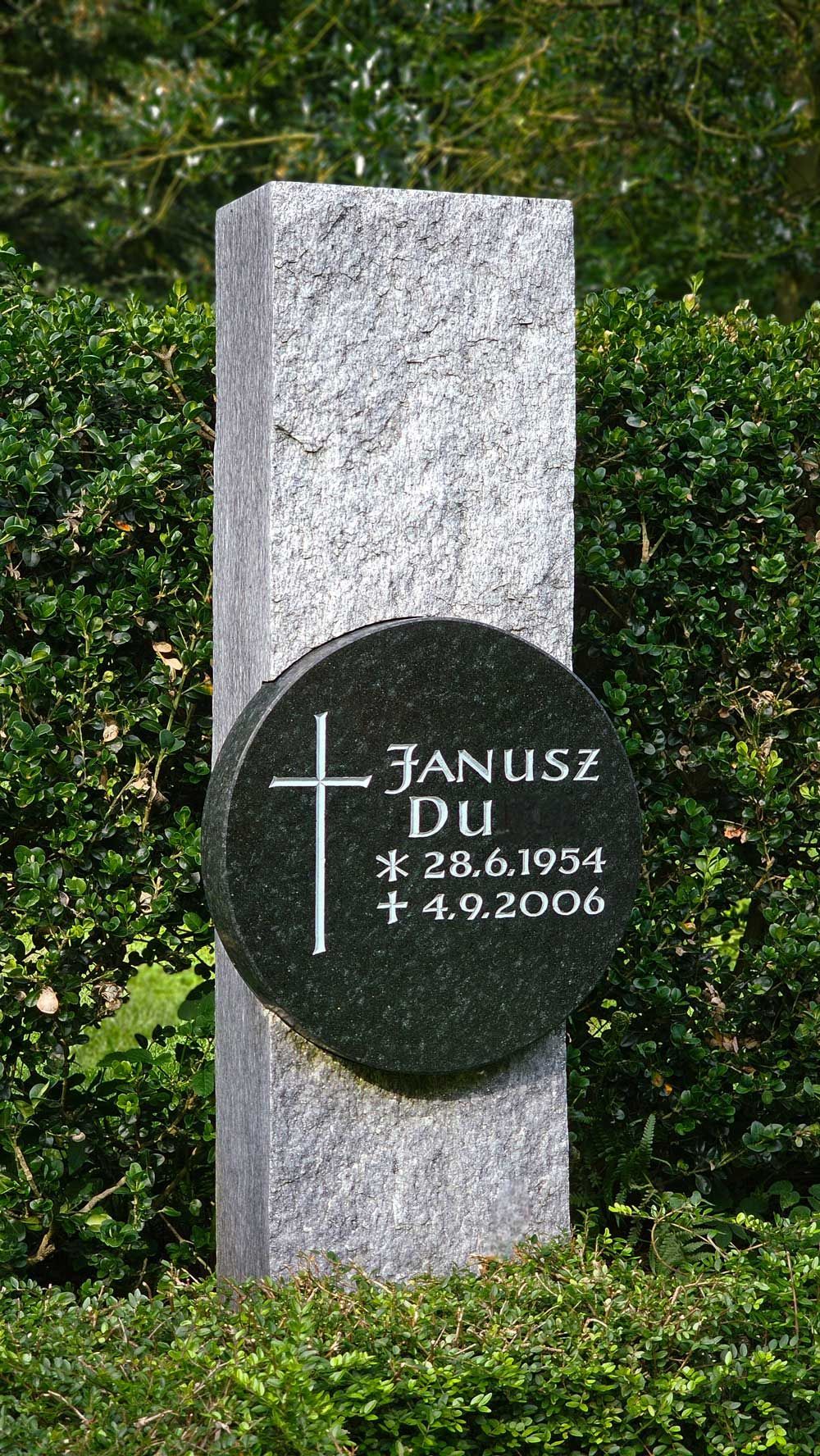 Grabstein von Janusz Du, ein dunkler runder Stein mit einem Kreuz, eingefasst in einen hellen rechteckigen Granitpfosten.
