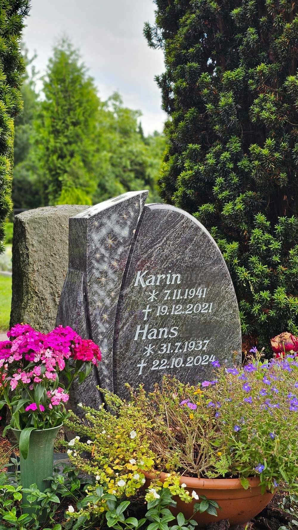 Grabstein mit den Namen „Karin“ und „Hans“ in einem Garten mit Blumen und Bäumen.