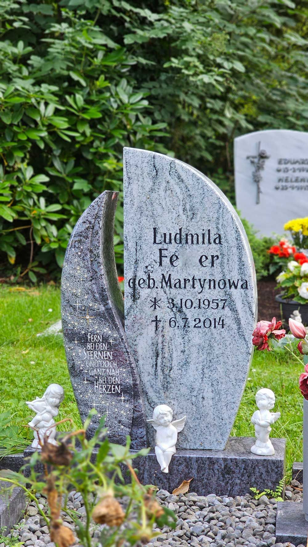 Grabstein für Ludmila Fer, geboren 1957, gestorben 2014, mit Engelsstatuen in einem Garten.
