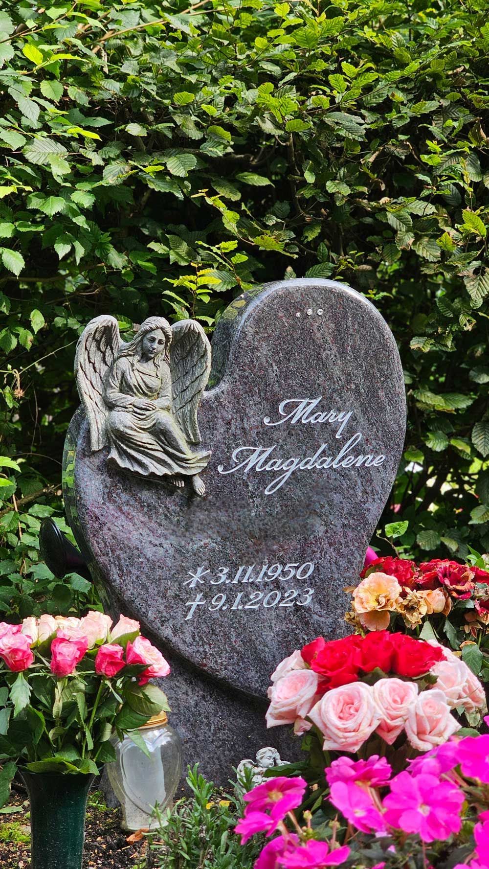 Herzförmiger Grabstein mit Engelsfigur, Inschrift und Blumen in einer Gartenlandschaft.