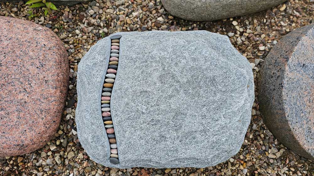 Grauer Fels mit einer senkrechten Reihe eingelassener kleiner Steine, umgeben von anderen Felsen auf Kies.