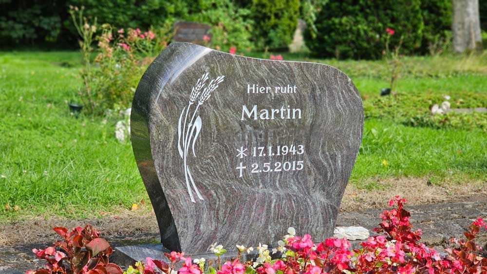 Grabstein für Martin mit den Daten 17.1.1943 - 2.3.2015, mit dekorativen Schnitzereien, in einer grasbewachsenen Umgebung mit Blumen.
