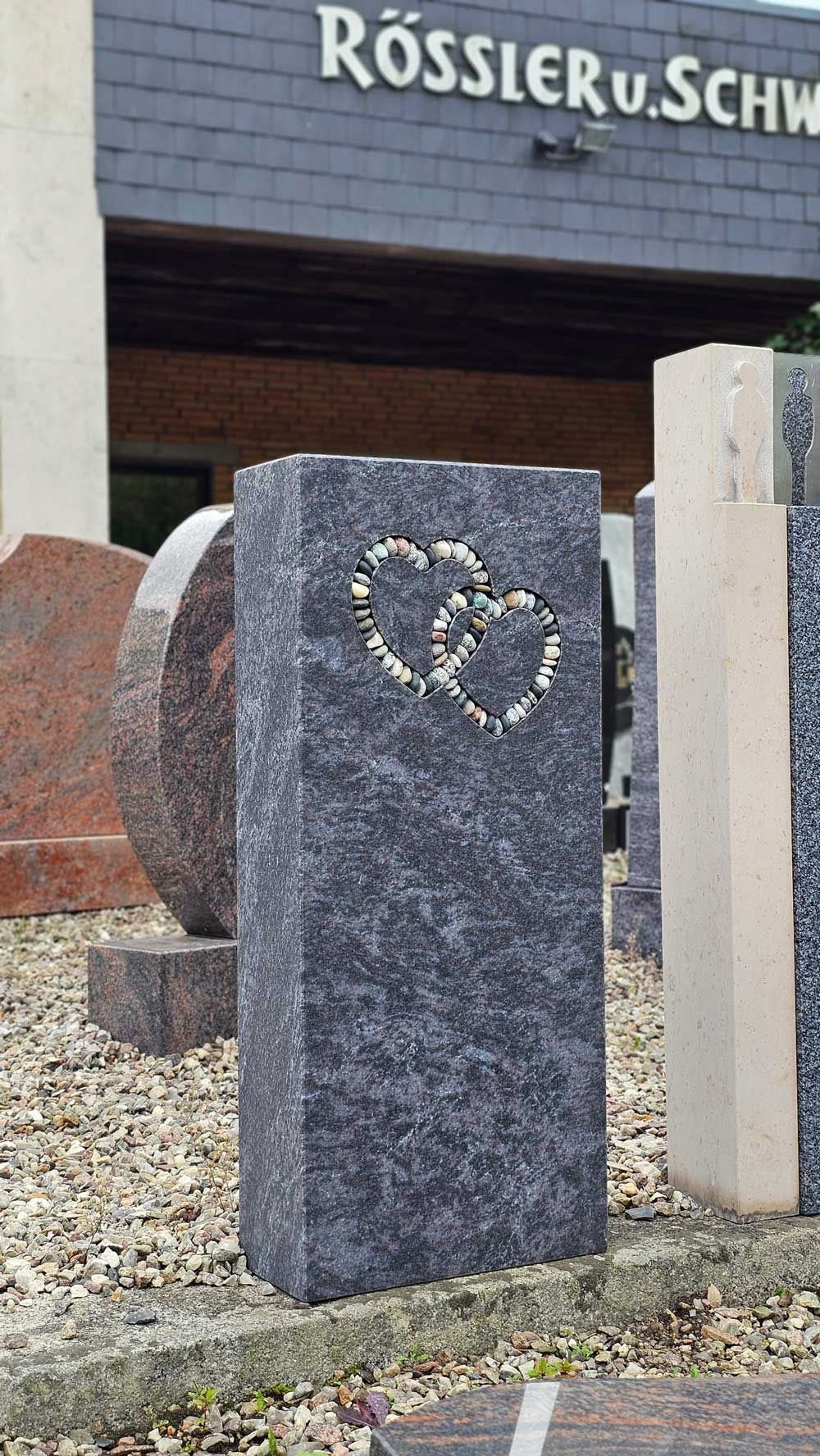 Grauer Granitgrabstein mit zwei Kieselherzen, vor einem Gebäude mit Text.
