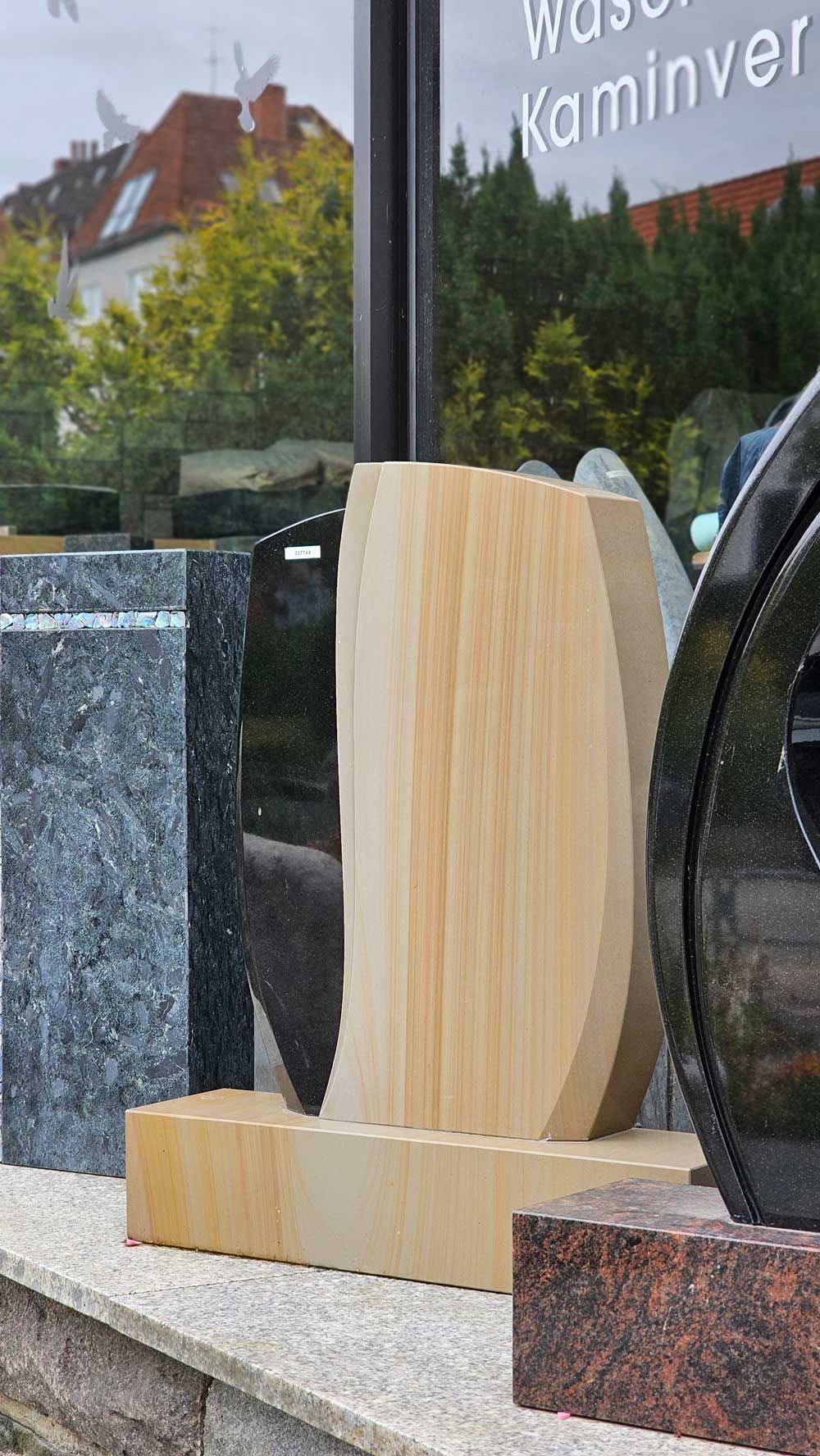 Vor einem Gebäude mit Glasfenstern sind Steindenkmäler in verschiedenen Formen und Farben aufgestellt.