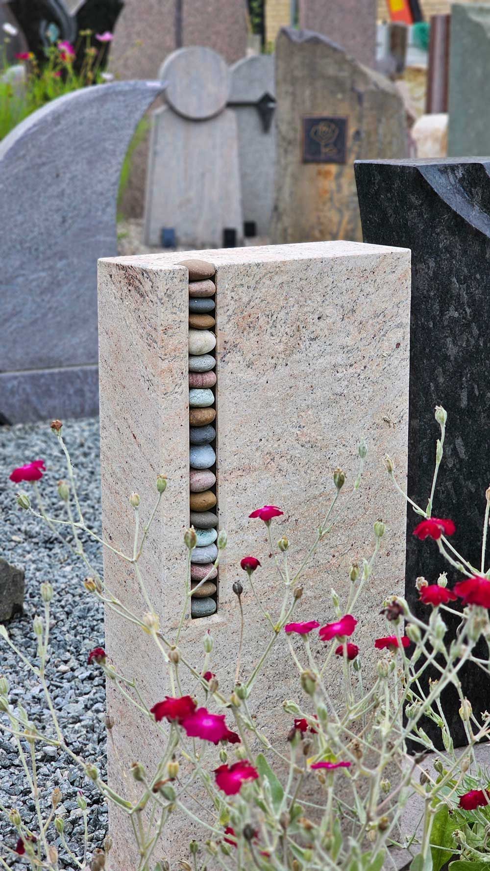 Steinsäule als Grabmarkierung mit in einer Nische gestapelten Kieselsteinen, umgeben von Blumen und anderen Grabsteinen.