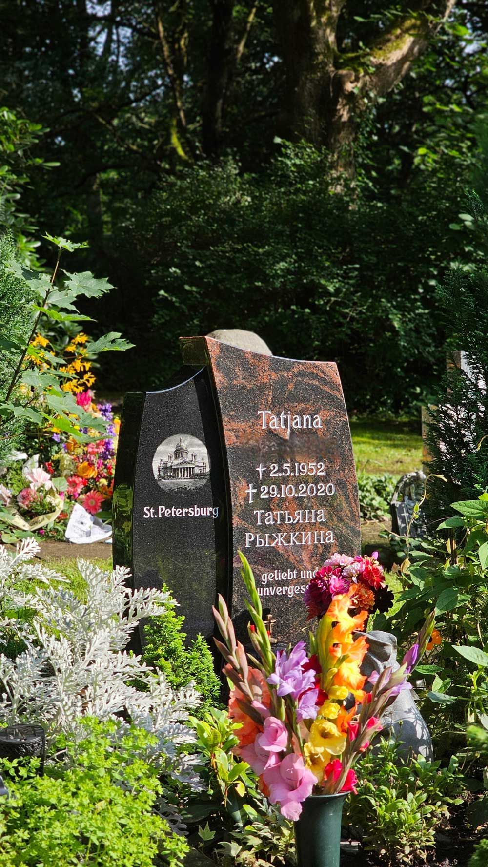 Grabstein in einem Garten. Schwarzer und rötlicher Granit, umgeben von Blumen und Grünpflanzen.