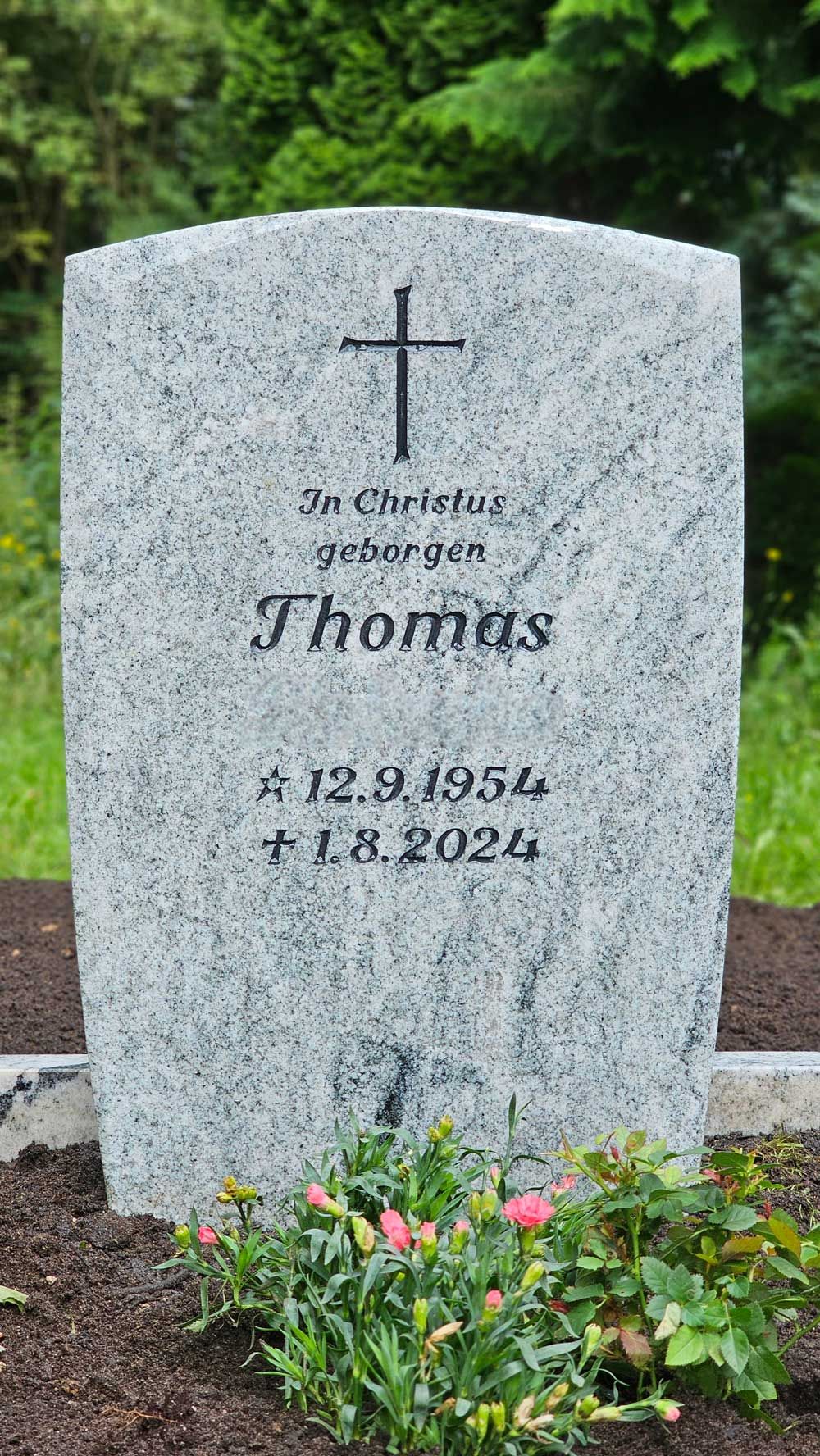 Grab von Thomas, Granitgrabstein mit Kreuz, Geburts-/Sterbedaten: 1954-2024, Blumen davor, im Freien.