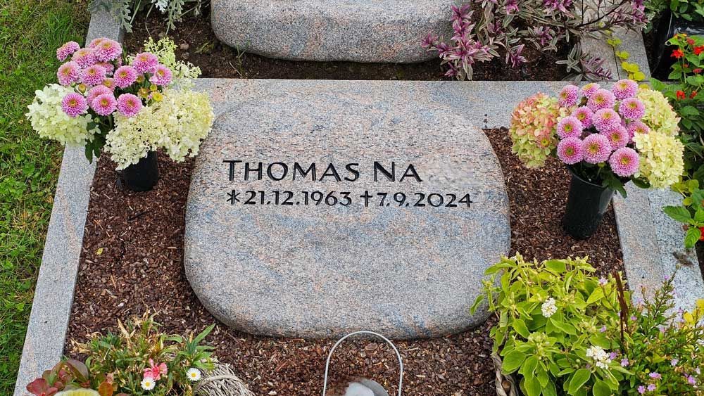 Grabstein mit Kreuz und Inschrift „Thomas“, Geburts- und Sterbedatum. Granit mit Blumen am Sockel.