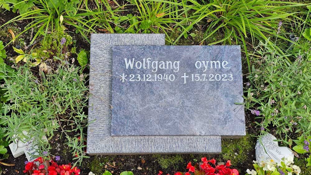 Grab mit Grabstein. Name „Wolfgang OyMe“ und Geburts-/Sterbedaten: 23.12.1940 – 11.1.2023. Umgeben von Grün und Blumen.
