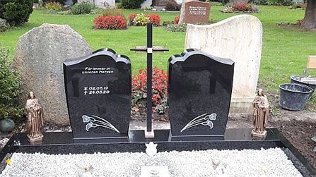 Zwei schwarze Grabsteine mit floralen Gravuren auf einer Grabstätte. Zwischen ihnen steht ein Kreuz.