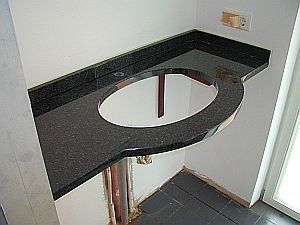 Schwarze Granitarbeitsplatte mit ovalem Spülbeckenausschnitt vor weißen Wänden. Darunter sind die Rohrleitungen sichtbar.