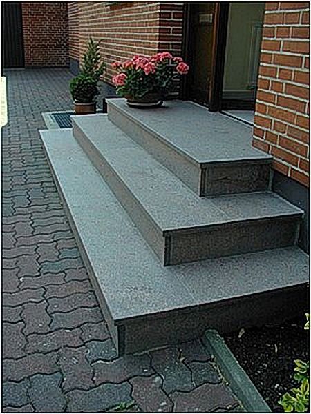 Drei Steinstufen führen zu einer Tür. Links ein Ziegelweg, auf der obersten Stufe Blumentöpfe, rechts eine Ziegelmauer.