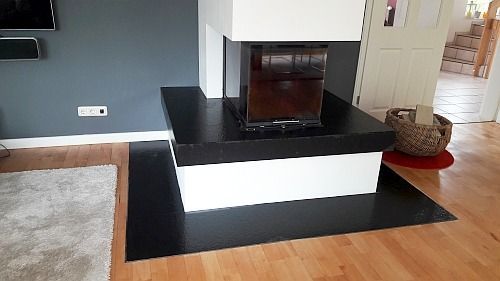 Moderner Kamin mit schwarzer Einfassung, weißem Sockel und Holzboden. Hinter dem Kamin ist eine graue Wand sichtbar.