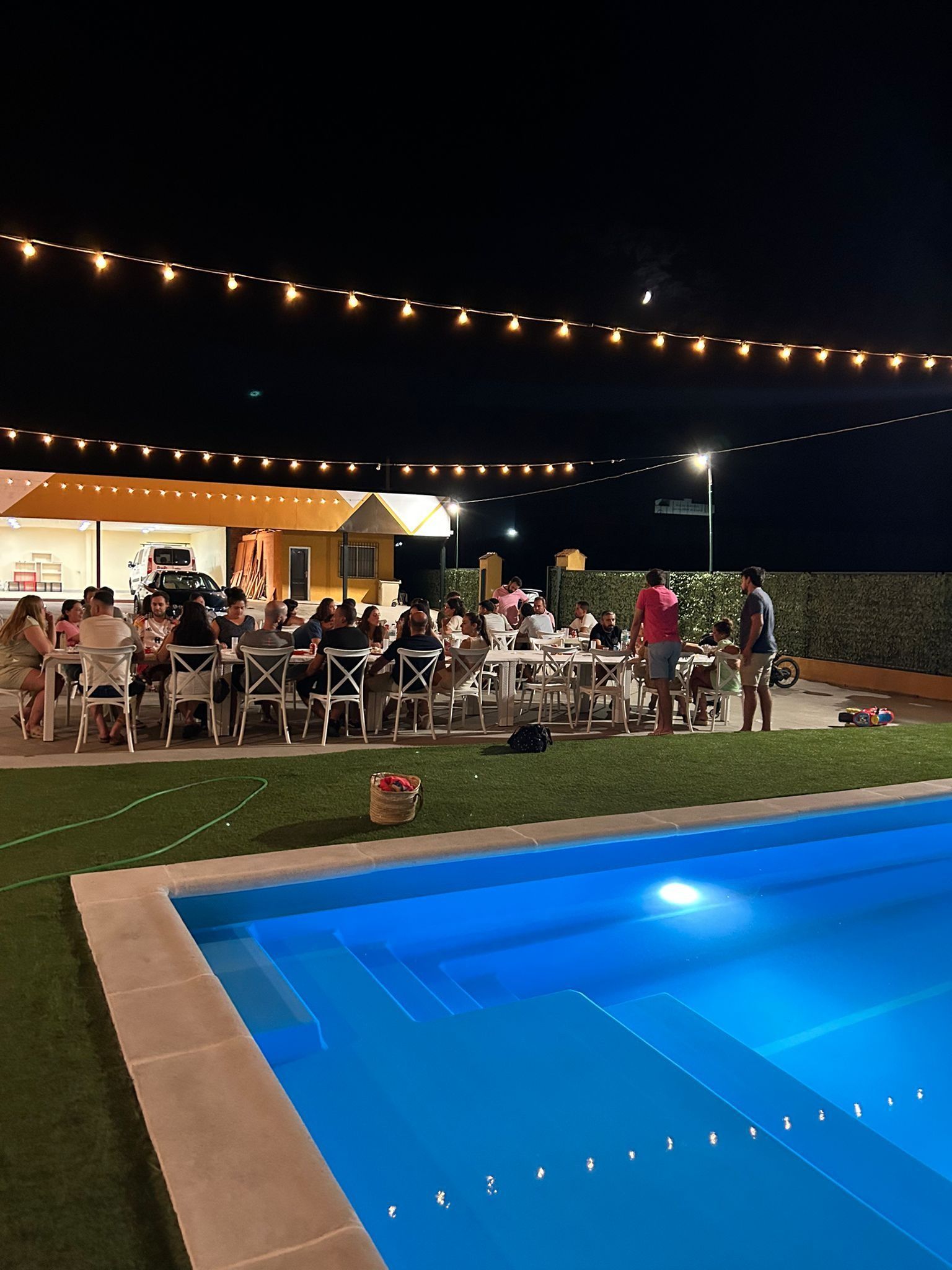 Un grupo de personas están sentadas en mesas junto a una piscina por la noche.