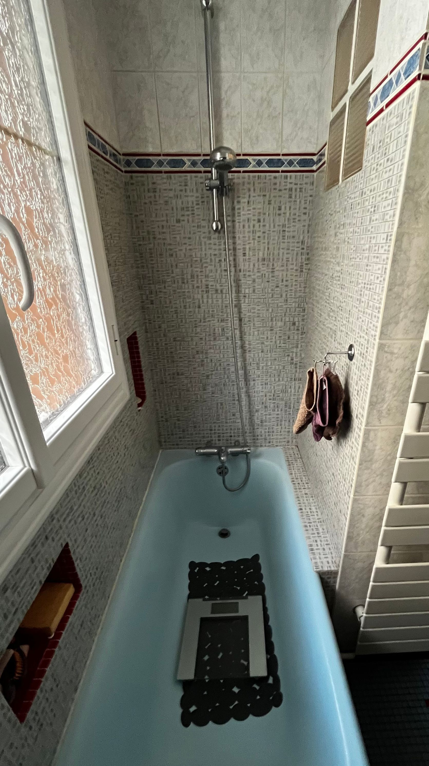 Baignoire bleue dans une salle de bain avec fenêtre, douche et pèse-personne. Le carrelage est blanc et bleu.