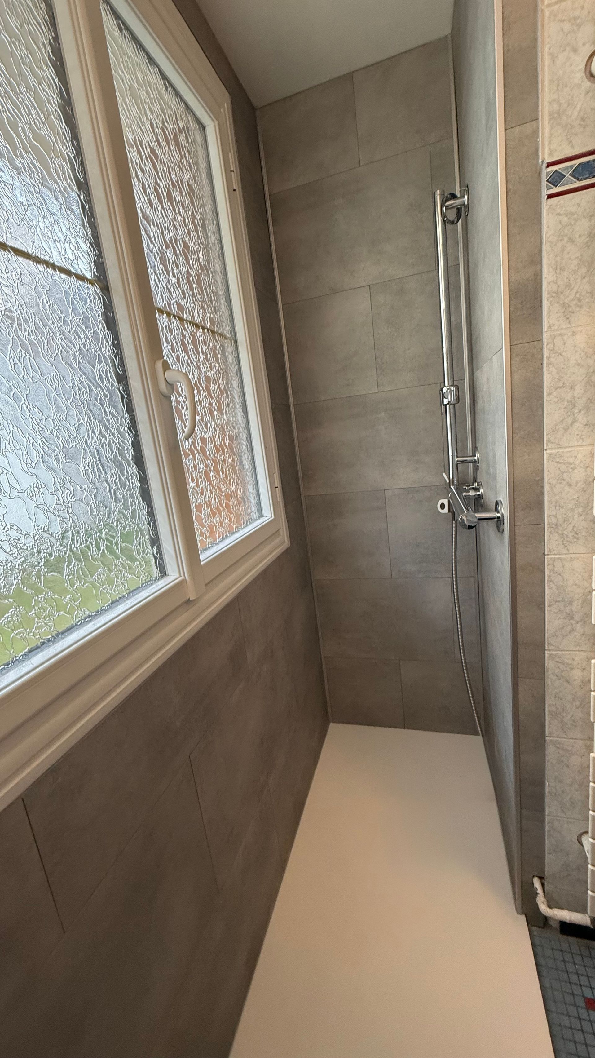 Douche avec fenêtre, carrelage gris et robinetterie chromée. Base et encadrement de fenêtre blancs.