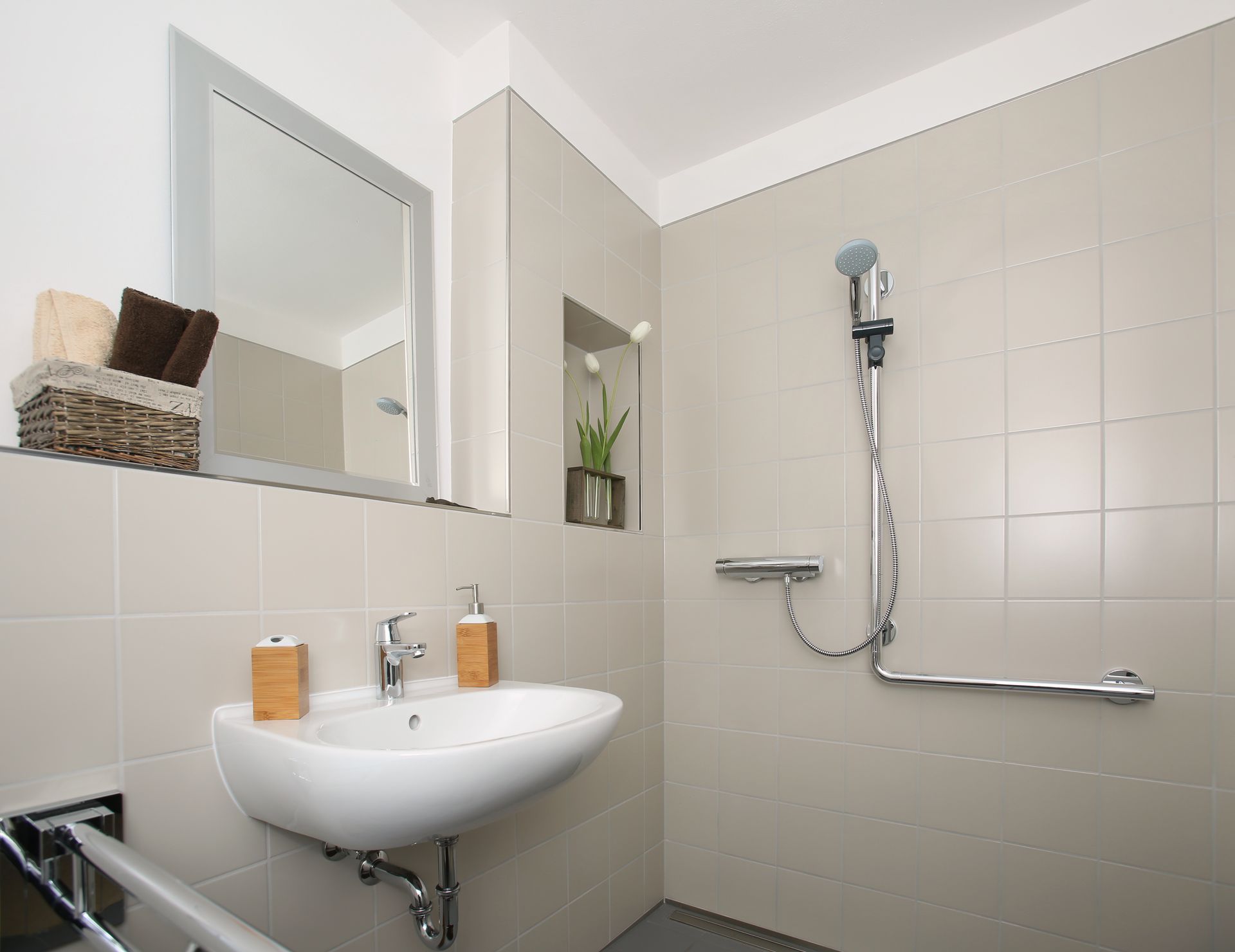 Salle de bain accessible avec lavabo mural, douche avec barre d'appui et carrelage de couleur neutre.