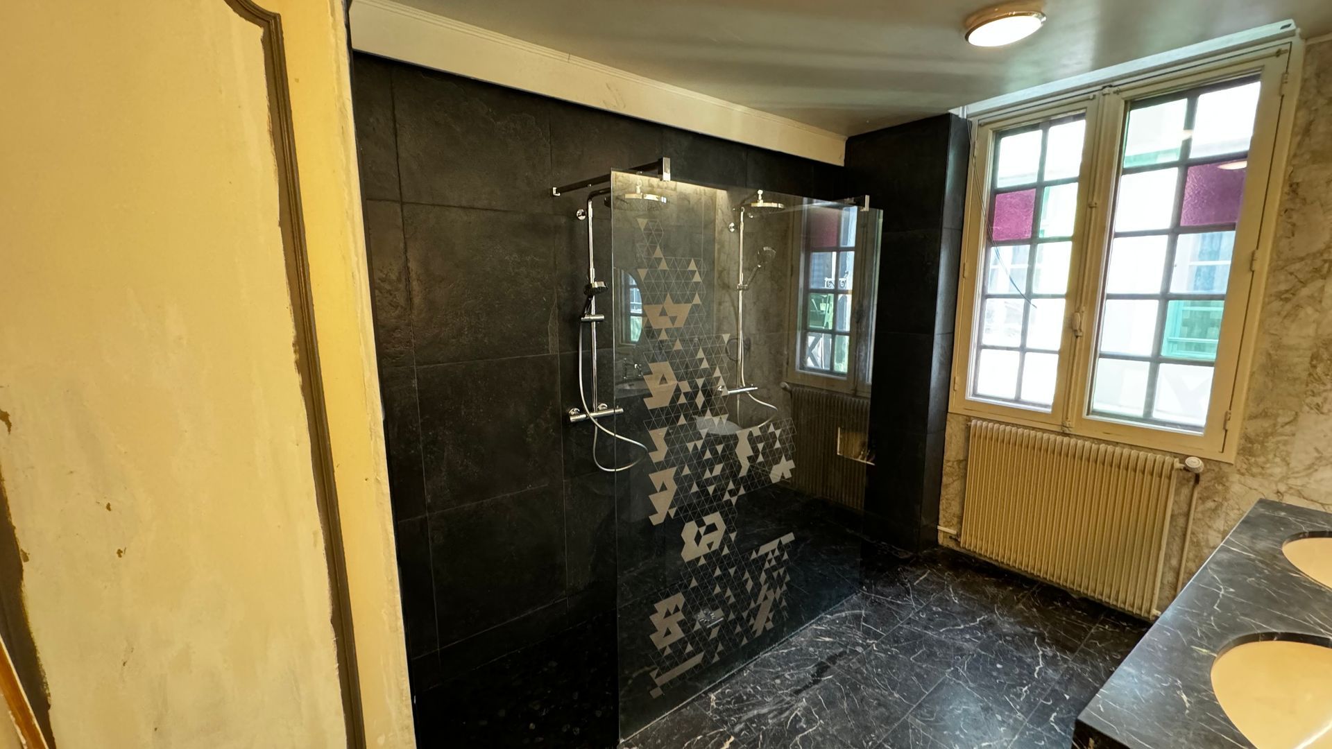 Salle de bain avec douche carrelée noire, portes vitrées et fenêtre aux accents violets.