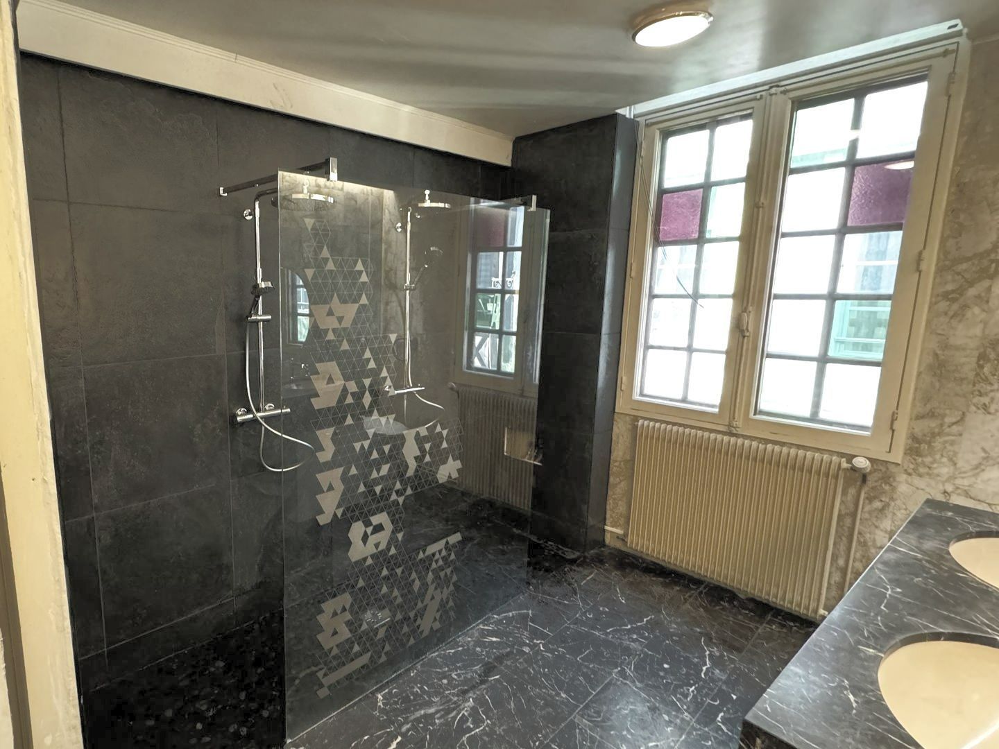 Salle de bain carrelée noire avec douche, fenêtre et lavabo. Sol effet marbre, murs sombres et lumière naturelle.