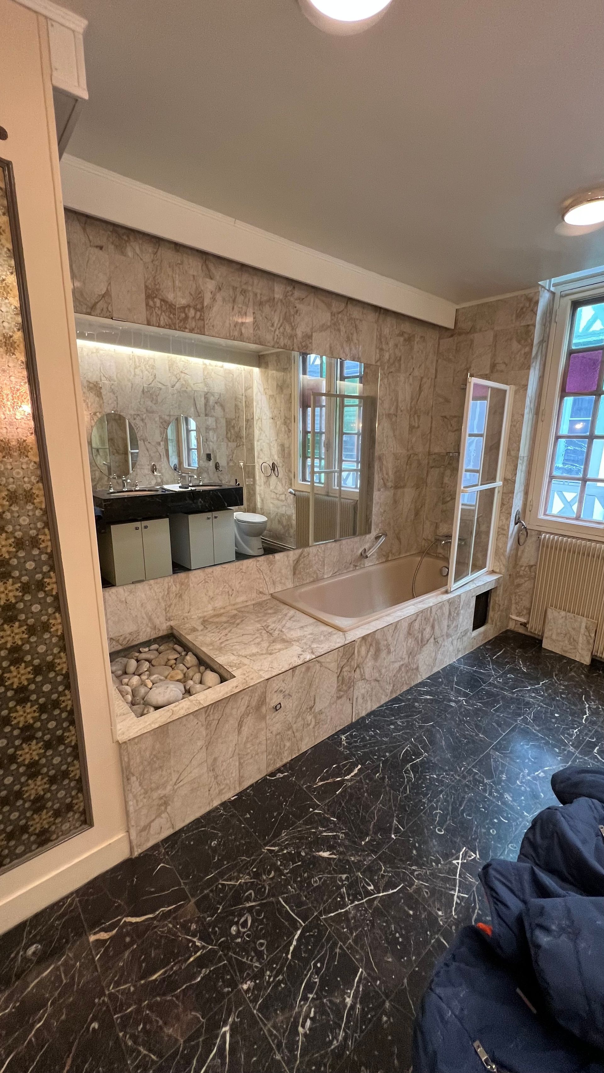 Salle de bain avec murs en marbre, baignoire encastrée, sol moucheté noir et grande fenêtre.