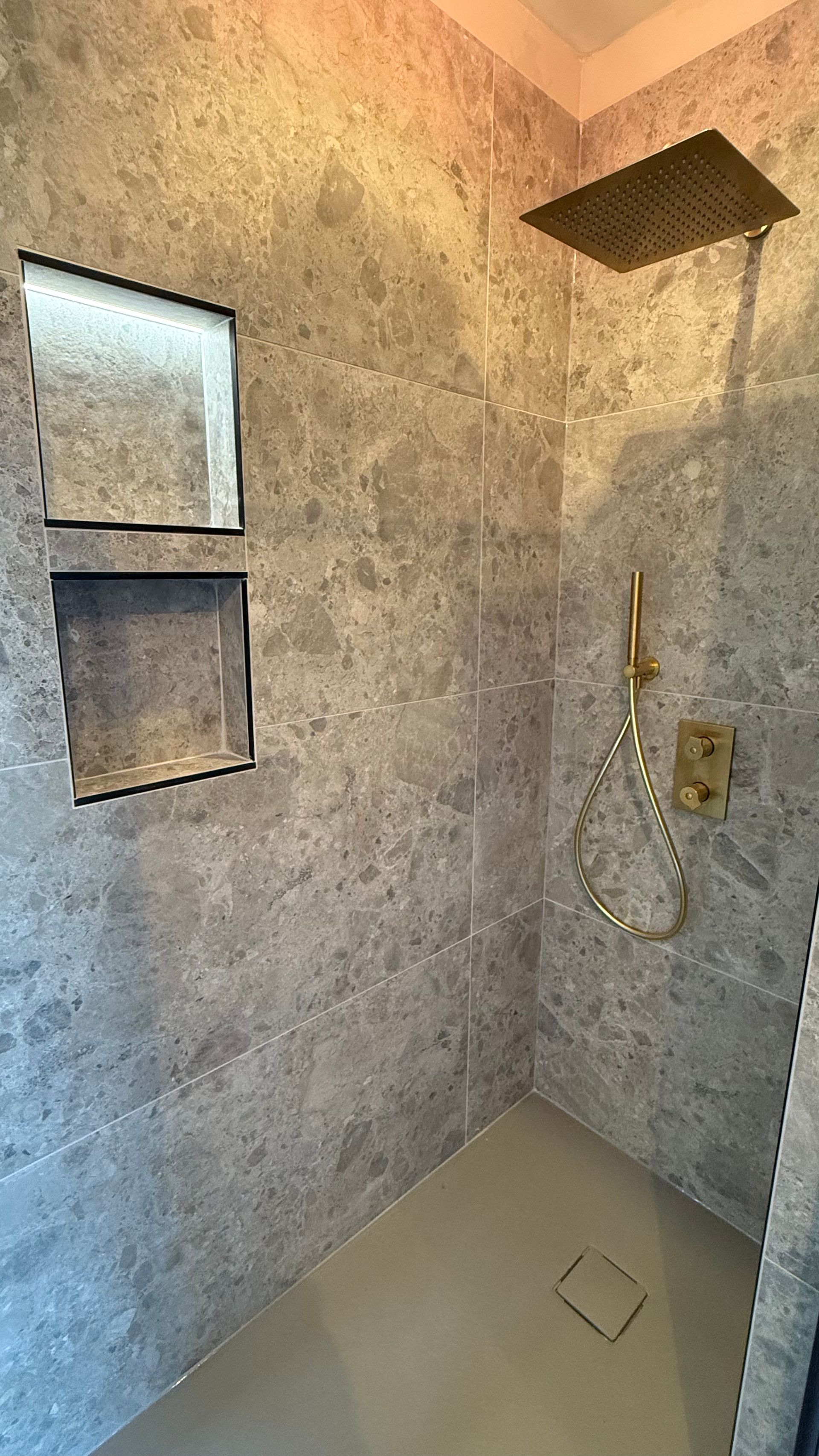 Douche carrelée grise avec accessoires dorés, étagères intégrées et pomme de douche à effet pluie.