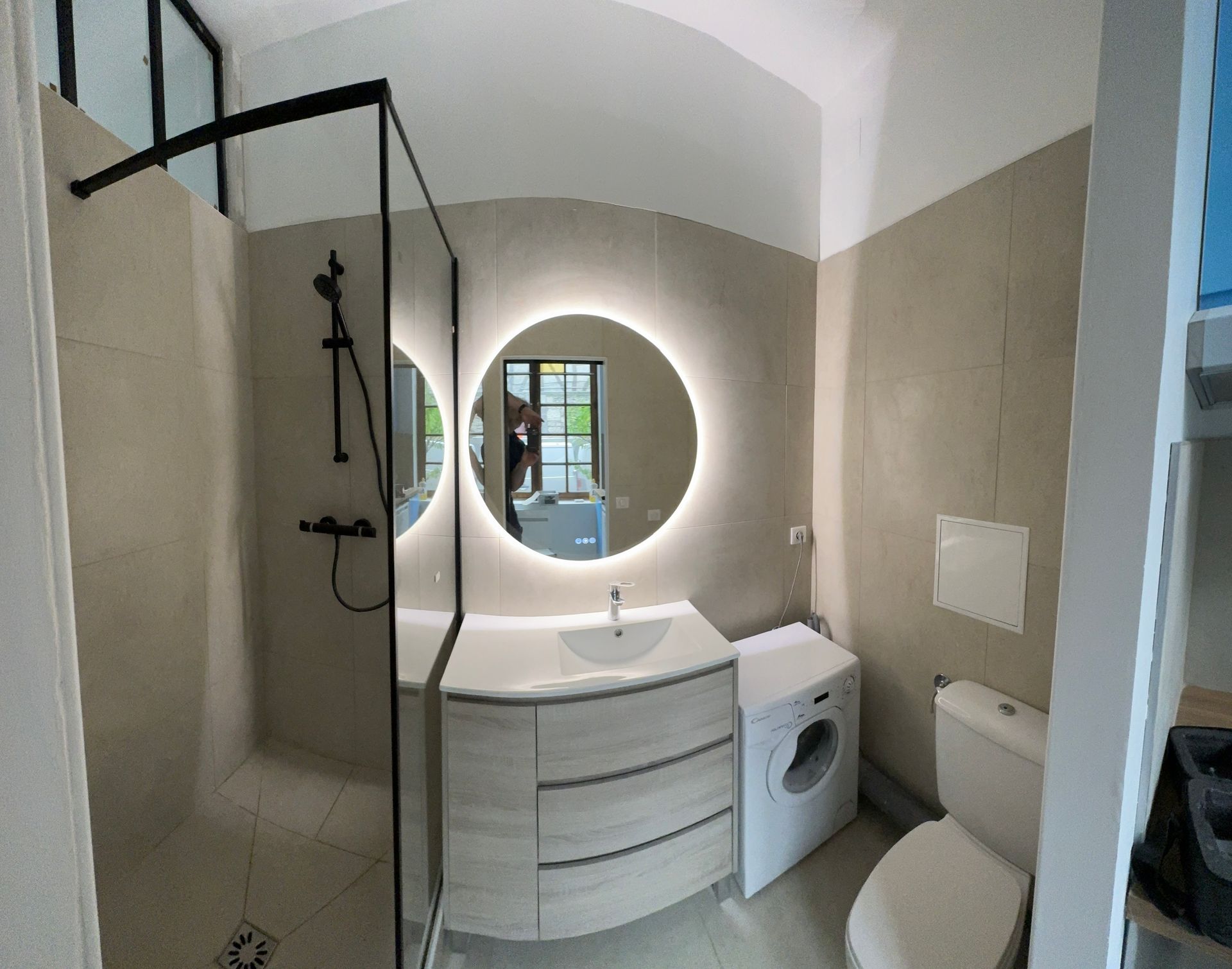 Salle de bain avec douche, miroir rond éclairé, meuble-lavabo, toilettes et lave-linge. Tons neutres.
