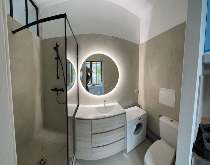 Salle de bain avec douche, miroir rond éclairé, meuble-lavabo, toilettes et lave-linge. Tons neutres.