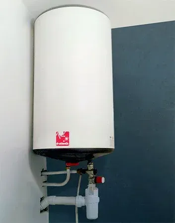 Chauffe-eau blanc monté sur un mur; comprend des tuyaux, des vannes et une étiquette rouge.