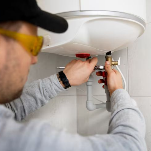 Une personne portant des lunettes de sécurité répare un chauffe-eau dans une salle de bain blanche.