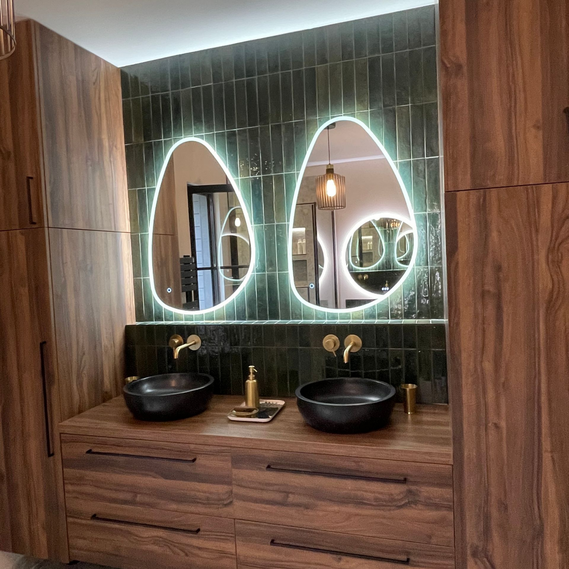 Salle de bain avec deux lavabos noirs, armoires en bois, carrelage vert et deux miroirs organiques éclairés.