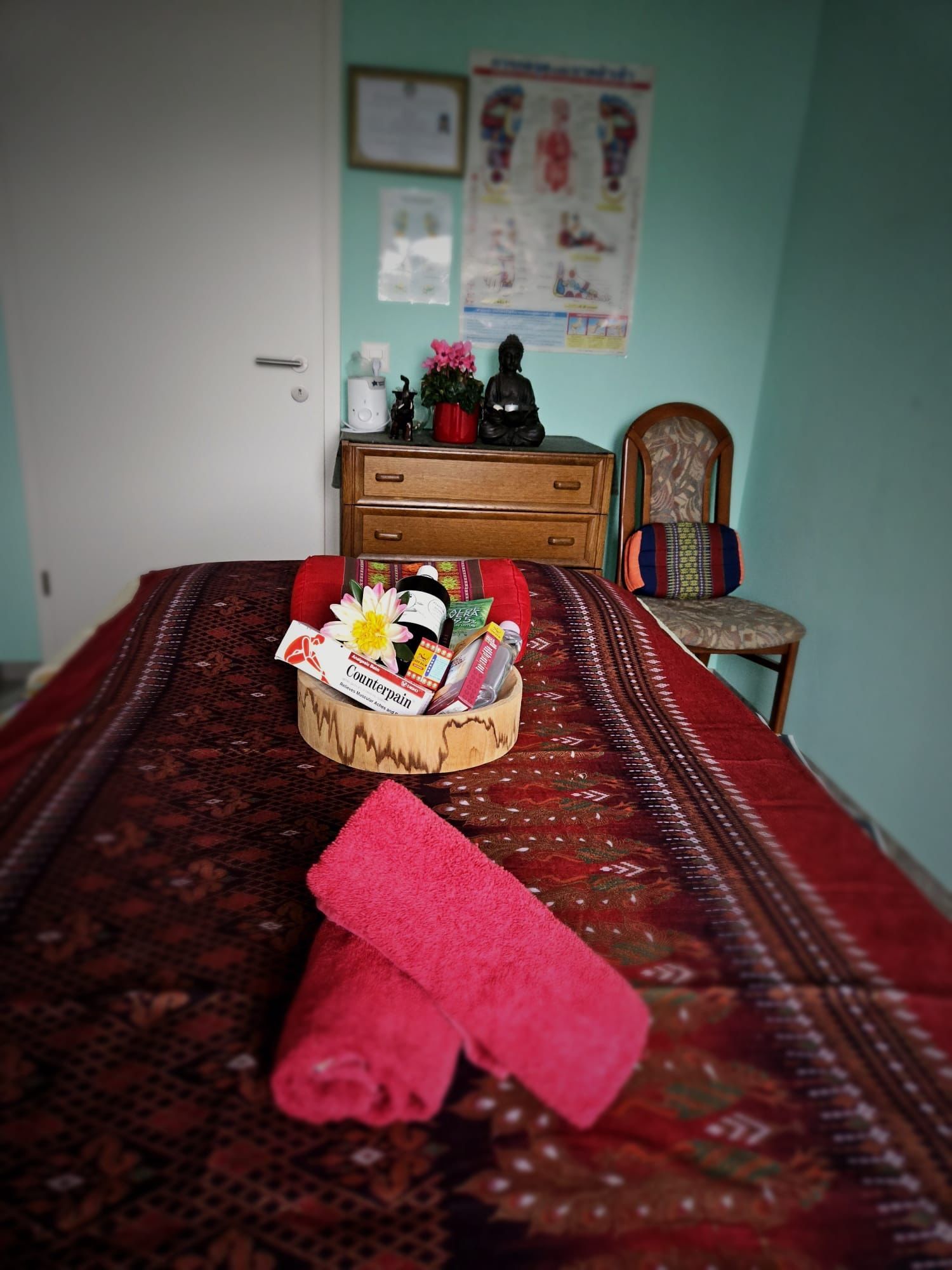 Massagetisch mit roten Handtüchern, Geschenkkorb, Kommode mit Dekorationen und Stuhl in einem Behandlungsraum.