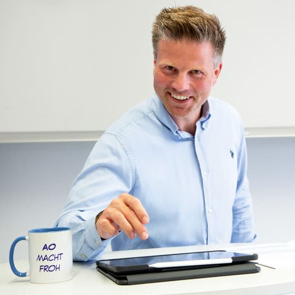 Ein Mann sitzt an einem Schreibtisch mit einer Tasse mit der Aufschrift „Ao macht froh“