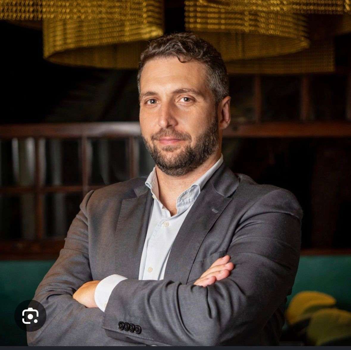 Hombre con chaqueta gris y brazos cruzados, posando en interiores con un fondo tenuemente iluminado.