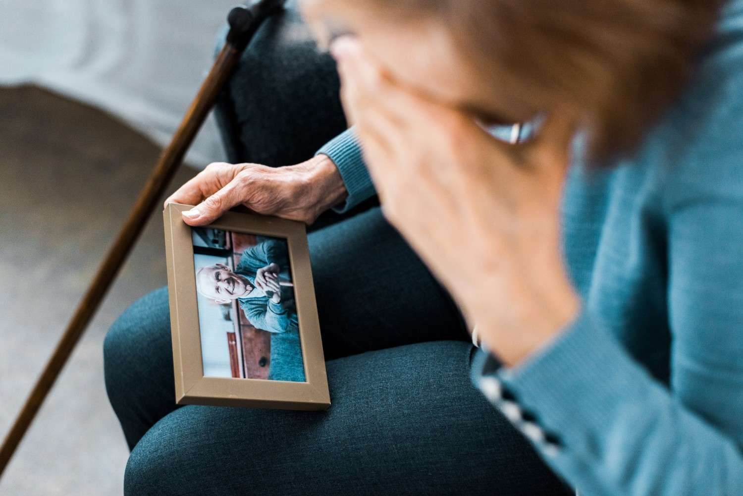 Mujer sosteniendo una foto enmarcada, llorando con la mano sobre la cara; sentada en un sofá.