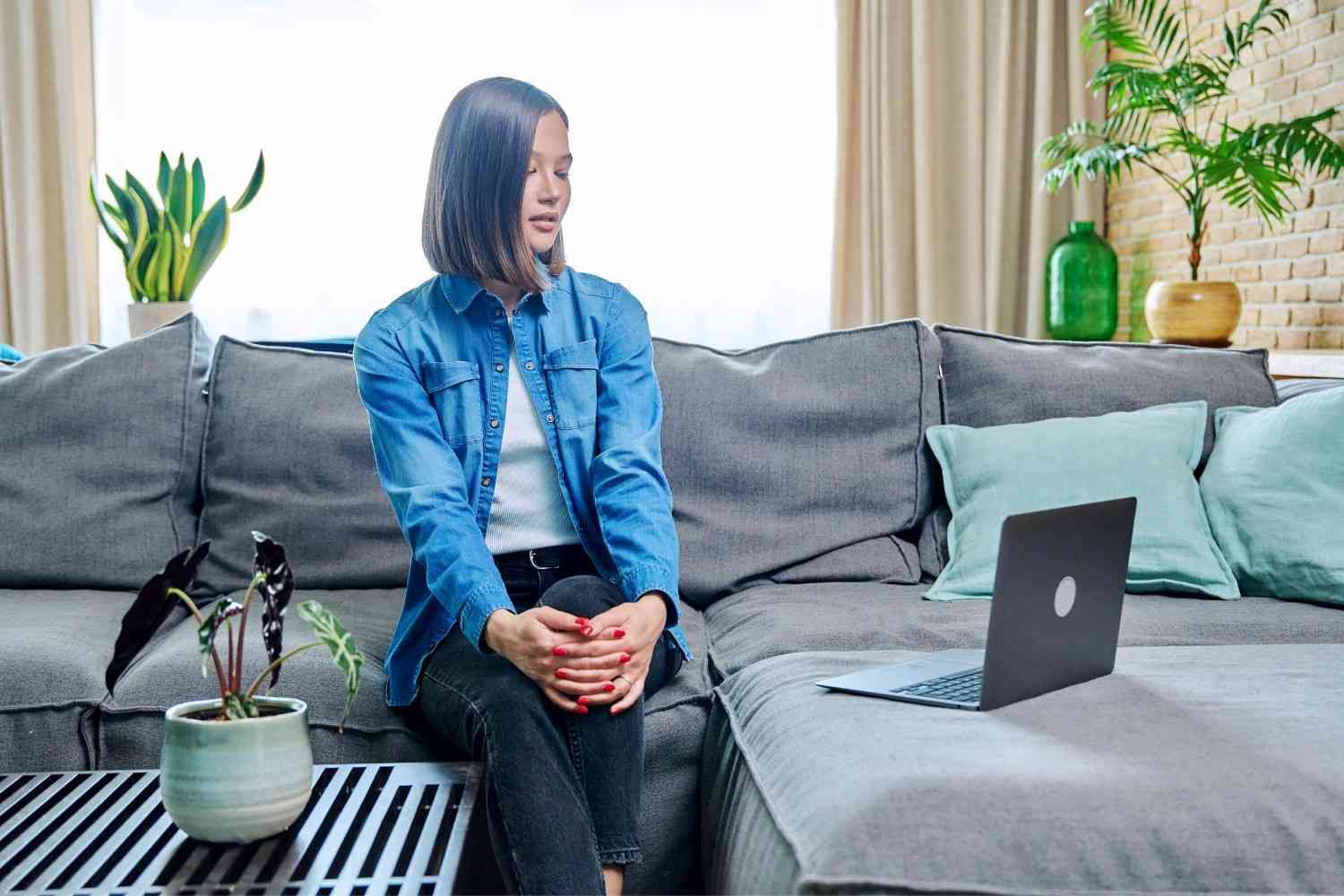 Persona en un sofá gris, mirando a otro lado, con un portátil cerca. Sala de estar con plantas.