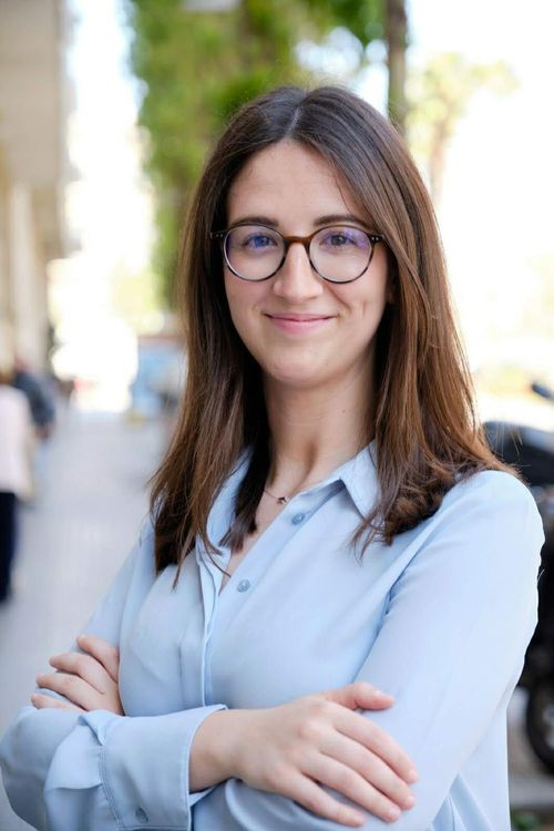 Mujer con gafas y brazos cruzados, sonriendo, frente a un fondo urbano borroso.