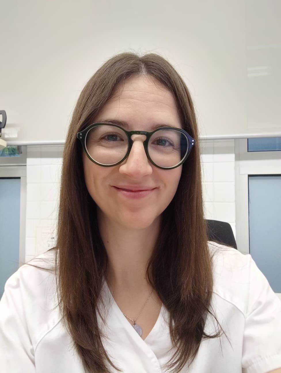 Mujer con cabello castaño y gafas sonriendo en el interior, vistiendo un uniforme blanco.