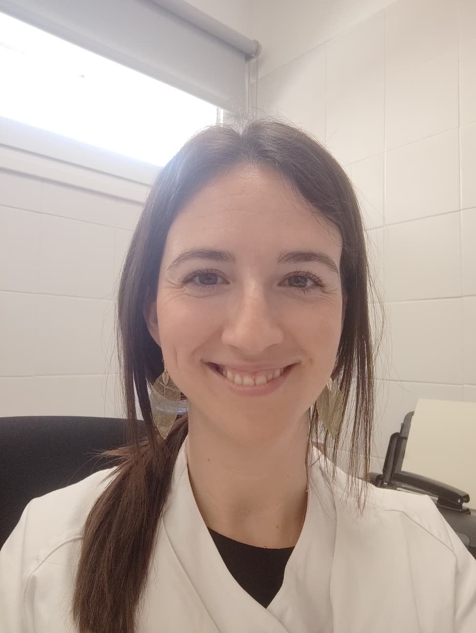 Una mujer con bata blanca sonríe, posiblemente en un entorno médico. Lleva el pelo oscuro recogido.