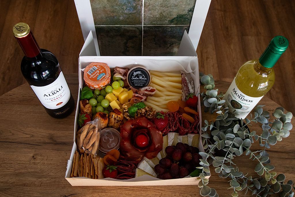 Caja de charcutería con botellas de vino y vegetación sobre una superficie de madera.