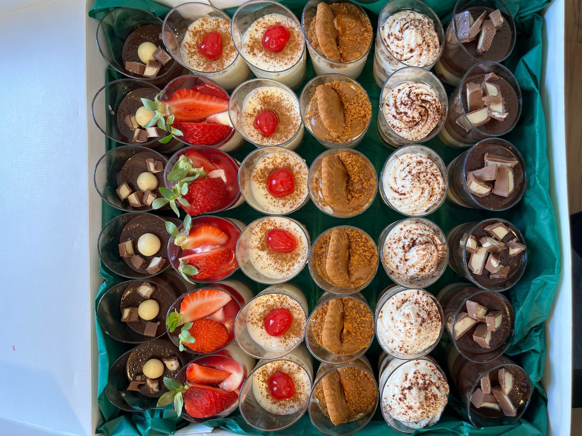 Una caja de postres variados en vasos transparentes: chocolate, fresas y delicias cubiertas con crema.