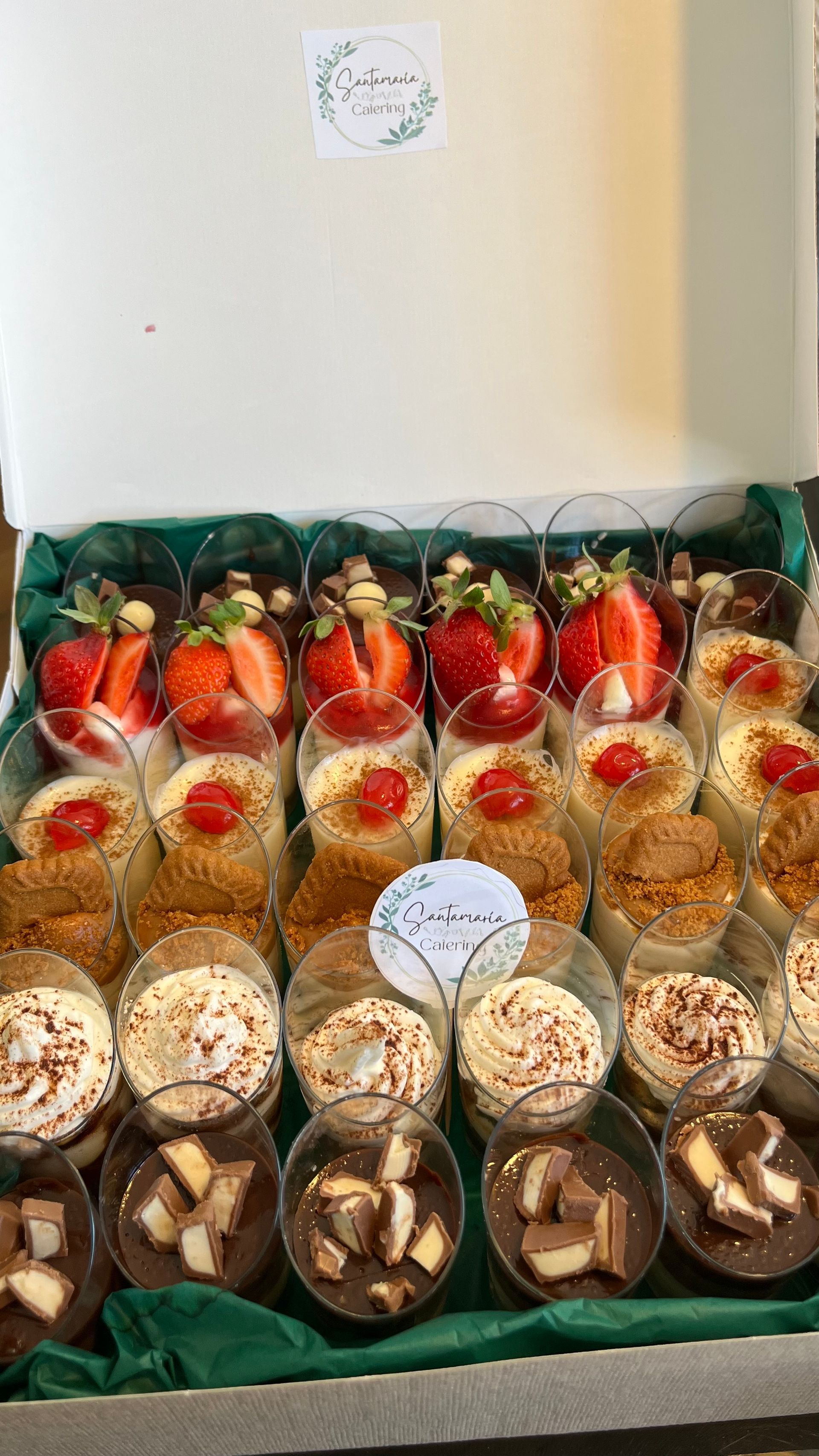 Caja de postres con diversas delicias: parfaits en capas, tazas de chocolate y coberturas de fresa, en una caja forrada de color verde.