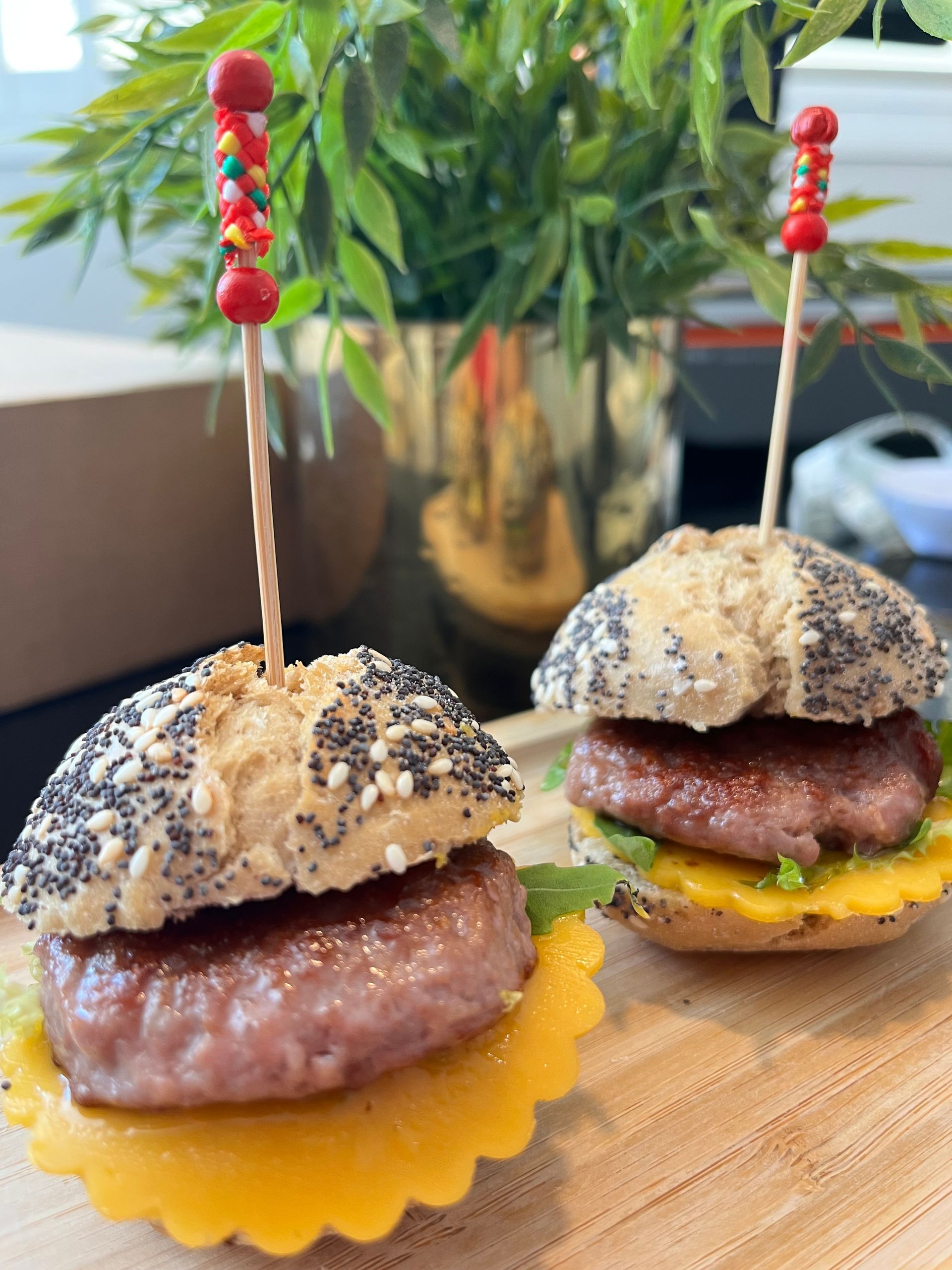Dos hamburguesas en miniatura sobre panecillos con semillas de amapola, ensartadas sobre una tabla de madera.