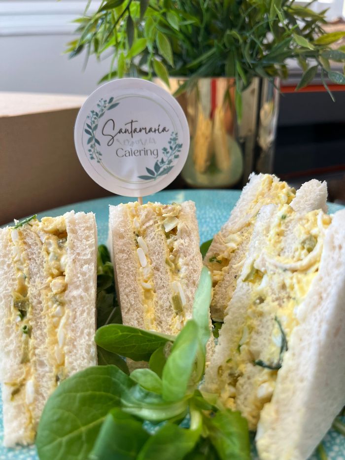 Sándwiches de ensalada de huevo en un plato azul, con vegetación y una maceta en tono dorado en el fondo.