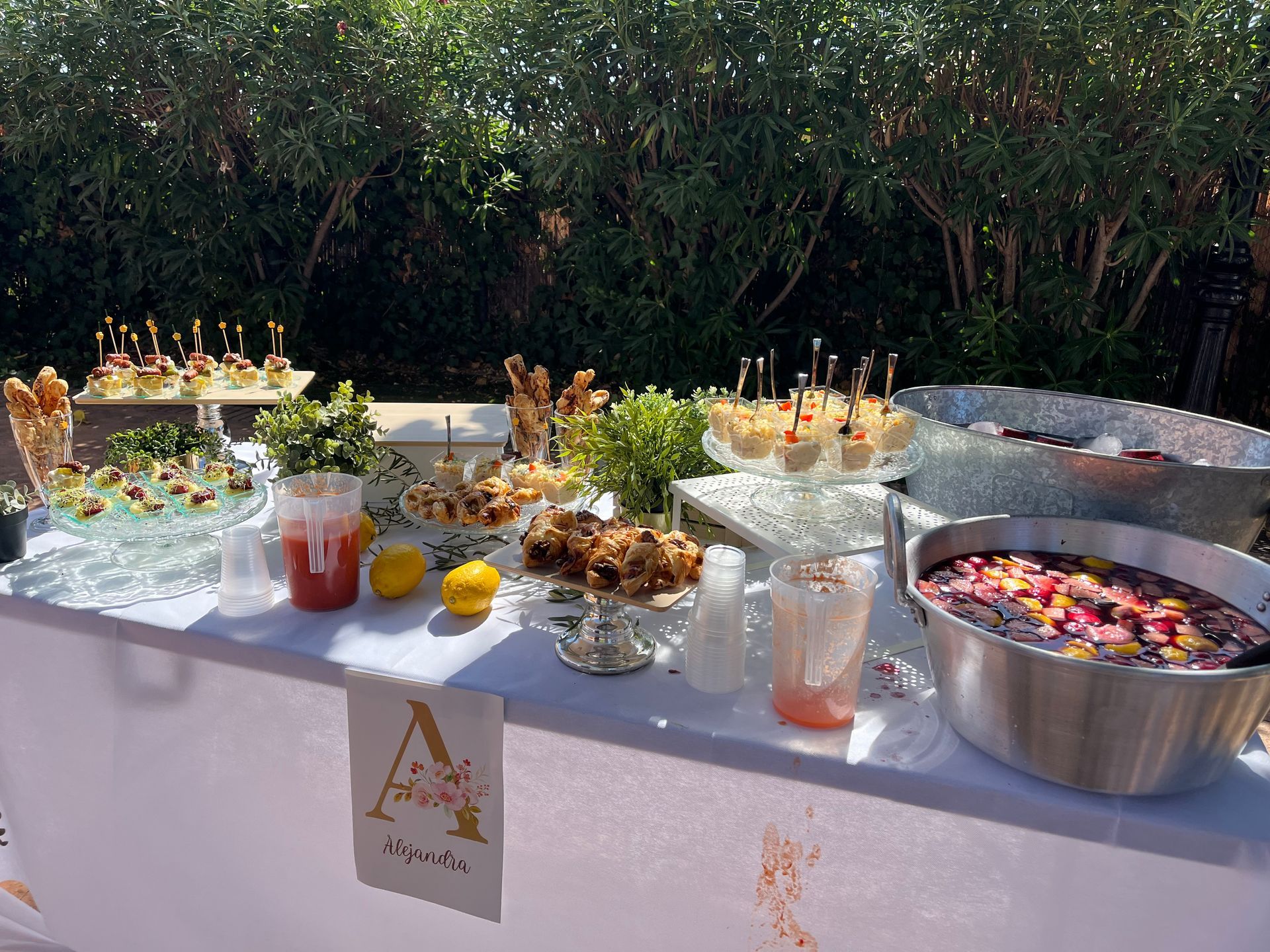 Una mesa con una variedad de aperitivos, bebidas y guarniciones, al aire libre, bajo el sol.